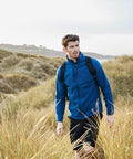 A person wearing a blue Craghoppers CR309 jacket and a backpack walks through tall grass in a hilly outdoor area.