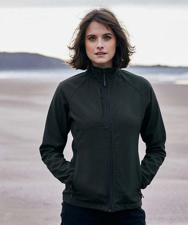 A person wearing a dark Craghoppers CR310 jacket stands on a beach with their hands in their pockets. The background features a shoreline and distant hills under an overcast sky.