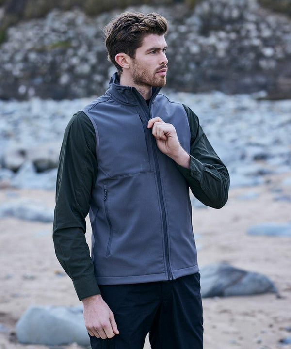 A man wearing a gray Craghoppers CR313 zip-up vest and dark long-sleeve shirt stands on a rocky beach.