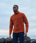 A person wearing an orange Craghoppers CR315 jacket and black pants stands near a rocky shore under a cloudy sky.