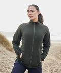 A person stands on a beach wearing a dark green Craghoppers CR316 zip-up jacket and dark pants, with hands in pockets and looking into the distance.