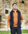 A man in an orange Craghoppers CR319 zip-up jacket and black vest stands in front of a stone wall, looking to the side with one hand in his pocket.