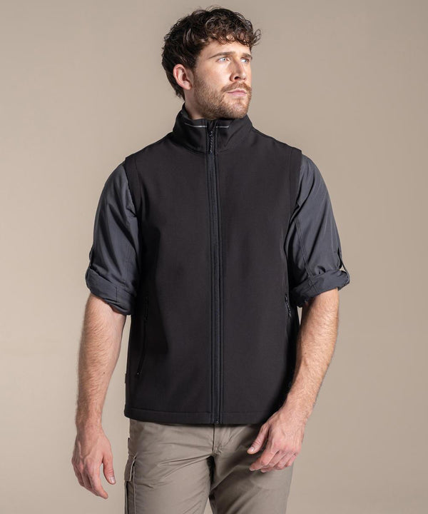 A man with short curly hair and a beard is wearing a Craghoppers Expert Basecamp II softshell gilet over a dark grey rolled-up sleeve shirt and light brown trousers, standing against a plain beige background.