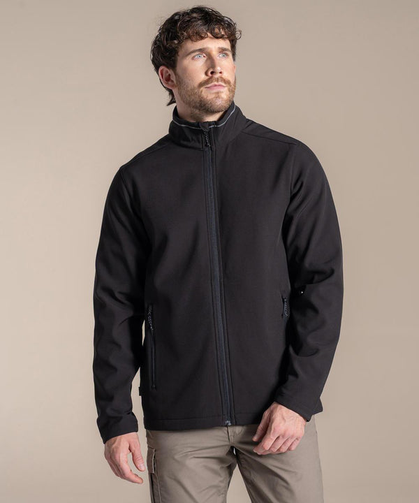 A man with short curly hair and a beard wears the Craghoppers Expert Basecamp II softshell jacket in black and light-coloured trousers, standing against a plain beige background while looking slightly upward to his right with a neutral expression.