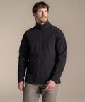 A man with short brown hair and a beard wears the Craghoppers Expert Kiwi 3-layer softshell jacket in black, paired with light grey trousers, standing against a plain beige background and gazing slightly up to his left.