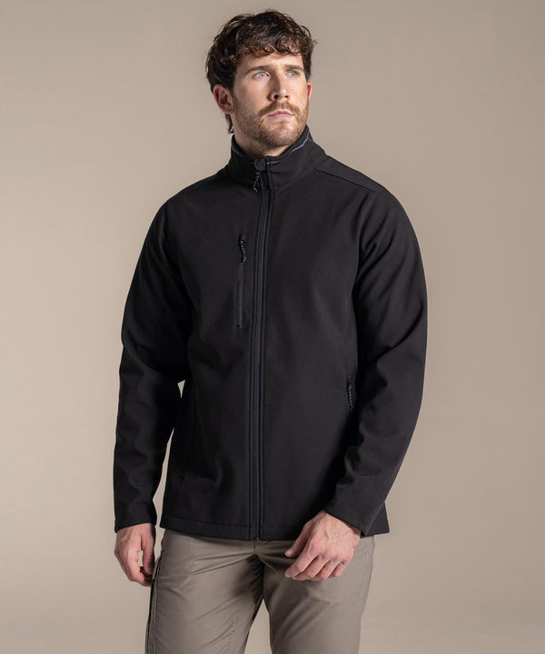 A man with short brown hair and a beard wears the Craghoppers Expert Kiwi 3-layer softshell jacket in black, paired with light grey trousers, standing against a plain beige background and gazing slightly up to his left.