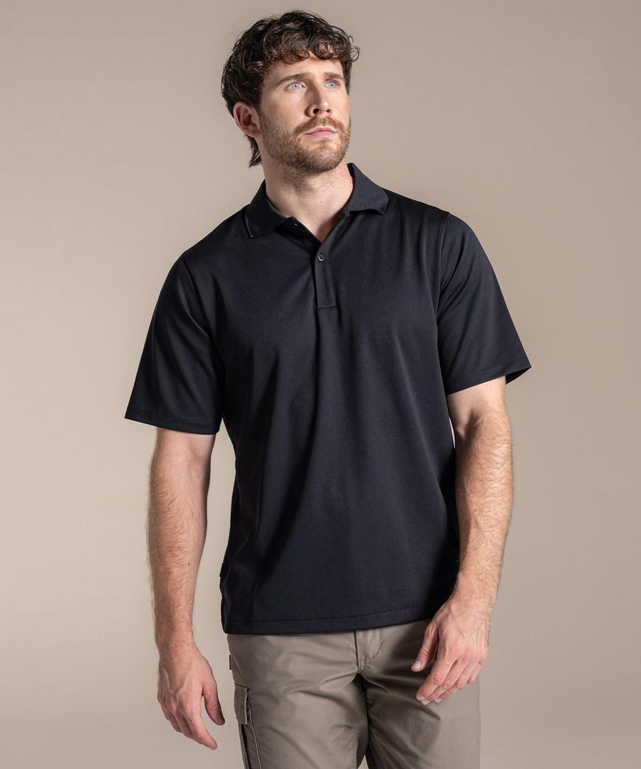 A man with wavy brown hair and a beard is wearing the Craghoppers Expert Basecamp wicking polo shirt in black, paired with grey trousers. He stands against a beige background, looking slightly upward to his left.