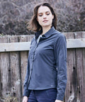 A person with shoulder-length hair is standing outdoors in front of a wooden fence, wearing a long-sleeved, button-up Craghoppers CR541 shirt and looking down to the side.
