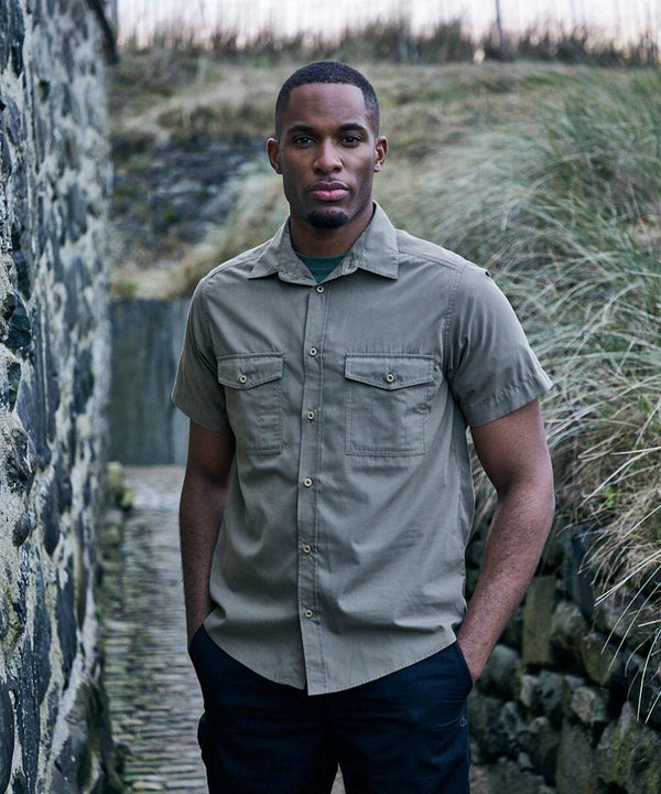 Man stands outside wearing a Craghoppers CR542 short-sleeved button-up shirt with hands in pockets. The background features a stone wall and tall grass.