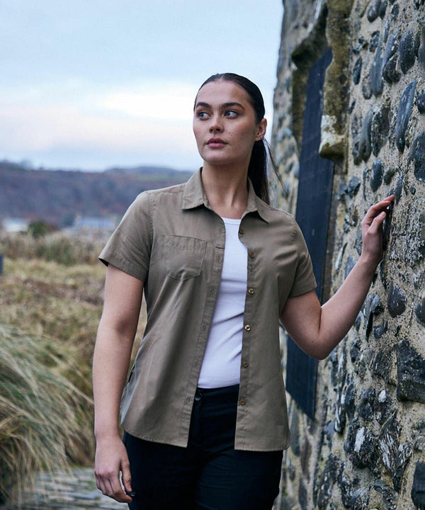 A person wearing a short-sleeve khaki CR543 by Craghoppers and black pants stands outdoors beside a stone wall, gazing into the distance.