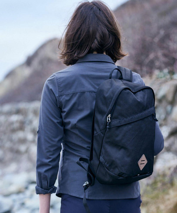 Rear view of a person with shoulder-length hair wearing a dark shirt and carrying a black CR622 backpack from Craghoppers, walking along a rocky terrain.