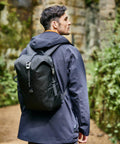 A person in a dark jacket and backpack stands on a dirt path, surrounded by greenery and rocks, looking ahead, equipped with the CR623 from Craghoppers.