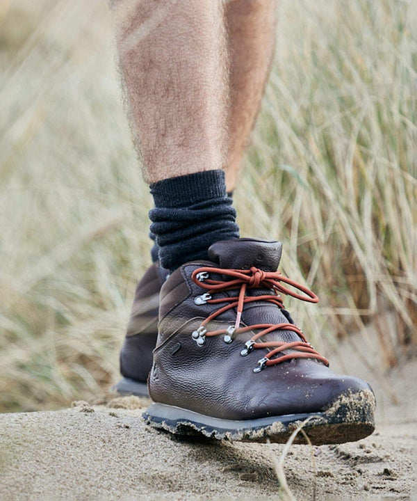 Close-up of a person wearing Craghoppers CR650 brown hiking boots with red laces, black socks, and standing on a sandy surface with dry grass in the background.