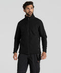A man with short curly hair and a beard is wearing a black Craghoppers CR702 jacket and black pants. He is standing against a light gray background.