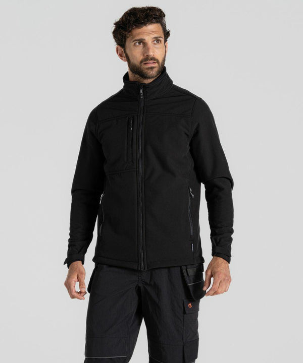 A man with short curly hair and a beard is wearing a black Craghoppers CR702 jacket and black pants. He is standing against a light gray background.