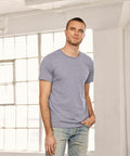 A man in a Bella + Canvas CV011 light gray t-shirt and jeans stands in front of large windows in a bright, industrial-style room.