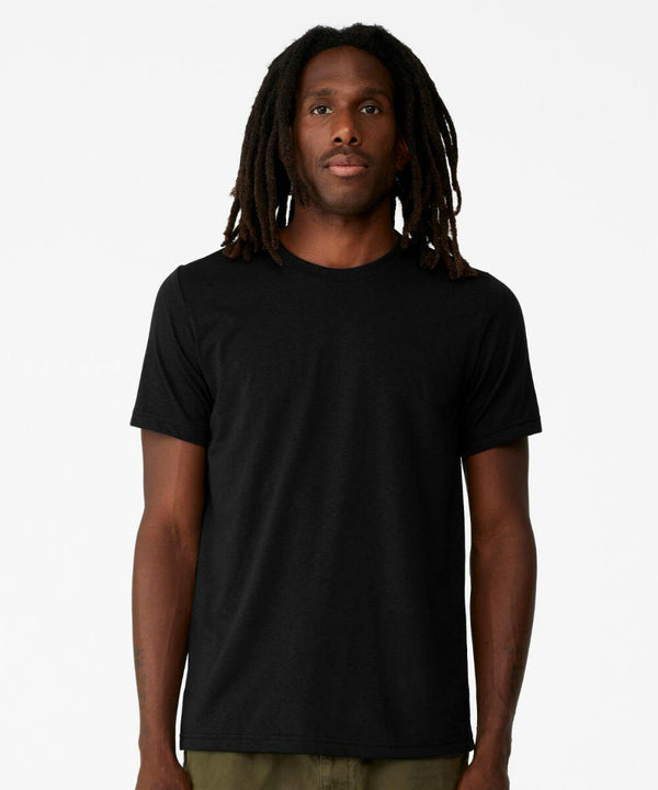 A person with long dreadlocks, wearing a plain black Bella Canvas CV012 and green pants, stands against a white background.