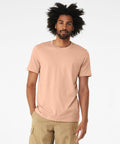 A man with an afro hairstyle is wearing a plain peach-colored Bella + Canvas CV01H and beige pants, standing against a solid white background.