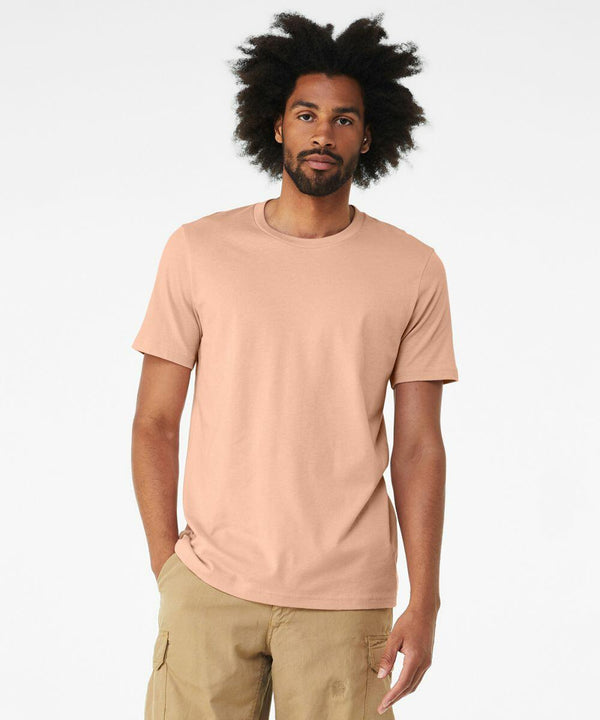 A man with an afro hairstyle is wearing a plain peach-colored Bella + Canvas CV01H and beige pants, standing against a solid white background.