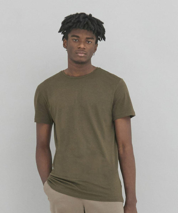 A person stands against a plain backdrop wearing a green EA002 by AWDis Ecologie T-shirt and beige pants, with one hand in their pocket.