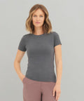 A woman with shoulder-length wavy hair is wearing an AWDis Ecologie EA01F gray short-sleeve t-shirt and pink pants. She is standing against a plain light background with one hand in her pocket.