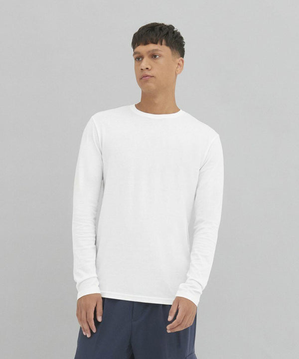 A person is wearing a plain white EA021 long-sleeve shirt by AWDis Ecologie and dark pants, standing with a neutral expression against a gray background.