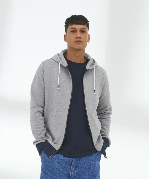 A person stands against a plain background, hands in pockets, wearing an AWDis Ecologie EA051 over a dark blue shirt and blue jeans.