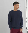 A person stands against a plain background, wearing a navy blue AWDis Ecologie EA062 knit sweater and red pants, looking to the right. Studio lighting equipment is partially visible on the right side.