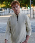 A young man with short curly red hair is wearing the Front Row Premium piqué 1/4-zip top in light beige, standing outdoors near trees and a road with green and red traffic lights blurred in the background.