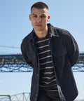 Man wearing a striped shirt and dark jacket standing outdoors with a bridge in the background, looking at the camera, showcasing the FR054 jacket by Front Row.