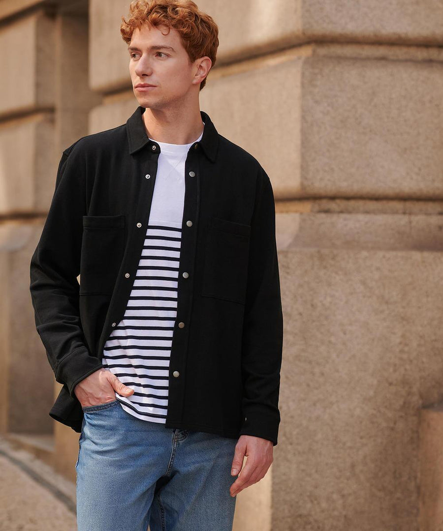 A man with short red hair stands outside by a stone building, wearing the Front Row Premium piqué overshirt over a white and black striped t-shirt and light blue jeans, one hand in his pocket.