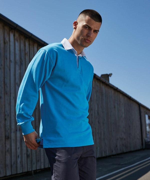 A person with a buzz cut stands outdoors wearing a blue long-sleeved Front Row FR100 shirt and navy pants, with a wooden building and clear sky in the background.