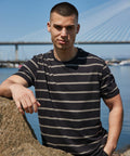 A man wearing a black and gray striped Front Row FR136 leans against a rock by the water, with a large bridge in the background.