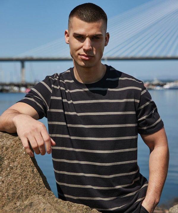 A man wearing a black and gray striped Front Row FR136 leans against a rock by the water, with a large bridge in the background.