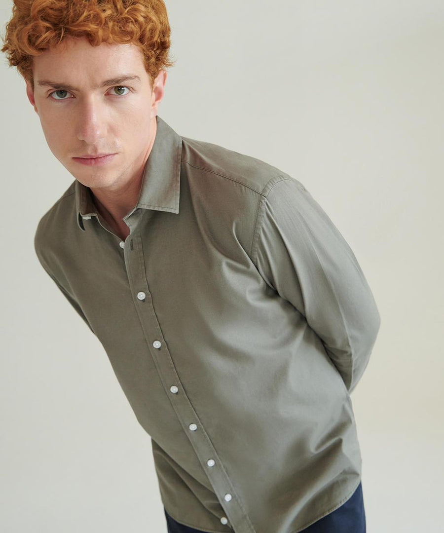 A man with short curly red hair, wearing a buttoned-up light grey Front Row Casual shirt, stands against a plain light background, leaning forward slightly with his hands behind his back.