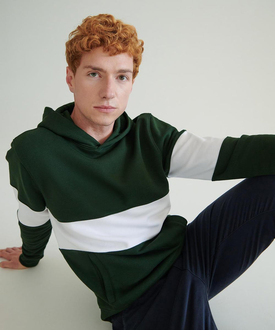 A person with short curly red hair wears the Front Row Heavyweight panelled hoodie in green with a white stripe and dark trousers, sitting on the floor with one arm extended back and a raised knee against a plain background.