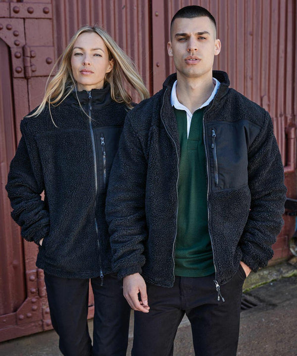 A man and woman stand side by side wearing Front Row FR854 black fleece jackets in front of a red, corrugated metal wall.