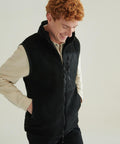 A person with curly red hair smiles while looking down, wearing a beige long-sleeve shirt and the Front Row Recycled Sherpa Gilet, which features zipped pockets, against a plain light background.