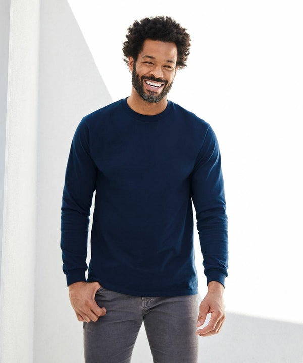 A person with curly hair wearing a GD004 by Gildan navy long-sleeve shirt and gray pants smiles while standing against a white background.