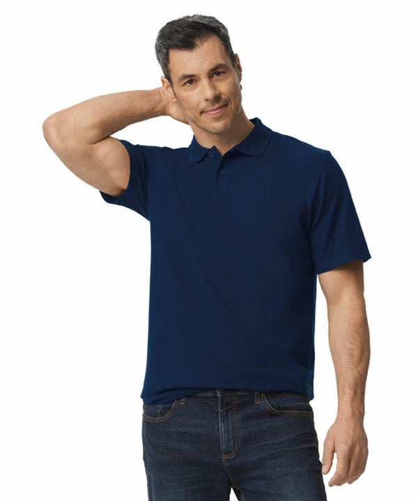 A man wearing a navy blue Gildan GD017 polo shirt and jeans stands with one hand behind his head and the other by his side.