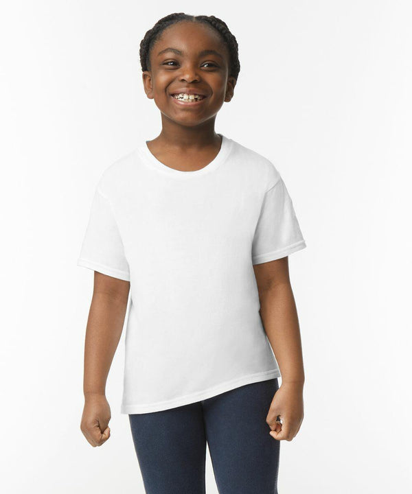 A young child smiles while standing, wearing a plain white Gildan GD01B t-shirt and dark pants. The background is plain and white.