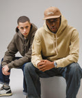 Two men wearing Gildan Hammer Maxweight adult hooded sweatshirts and jeans sit side by side. One wears a beige hoodie and Union cap, the other an olive hoodie. Both face the camera with neutral expressions.