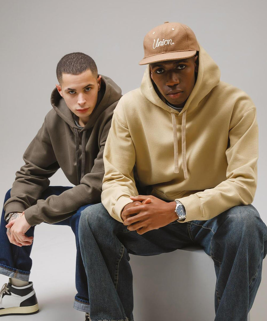 Two men wearing Gildan Hammer Maxweight adult hooded sweatshirts and jeans sit side by side. One wears a beige hoodie and Union cap, the other an olive hoodie. Both face the camera with neutral expressions.