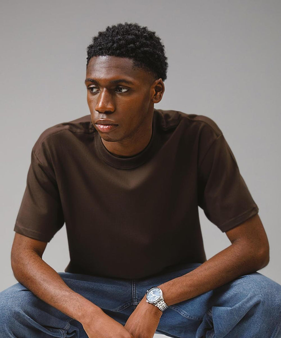 A young man with short curly hair, wearing a Gildan Hammer Maxweight adult t-shirt, blue jeans, and a silver wristwatch, sits against a plain grey background looking slightly to his left.
