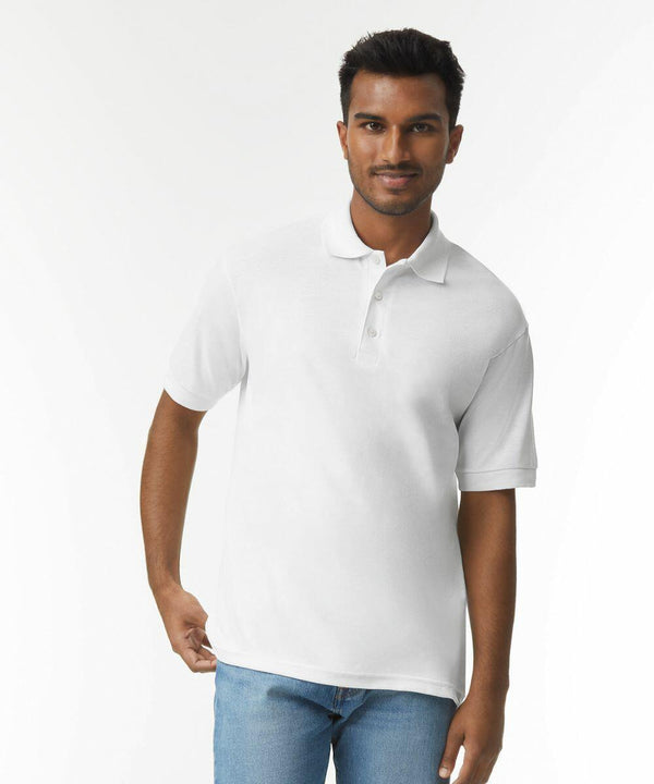 A man wearing a white Gildan GD040 polo shirt and blue jeans stands against a plain white background. He has short dark hair and a slight smile on his face.