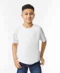 A young boy with short curly hair stands against a white background, wearing a plain white GD05B t-shirt by Gildan and blue jeans, with his right hand in his pocket.