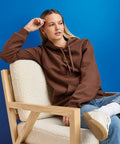 A person wearing a brown Gildan GD067 hoodie and blue jeans sits on a wooden chair with a white cushion against a blue background.
