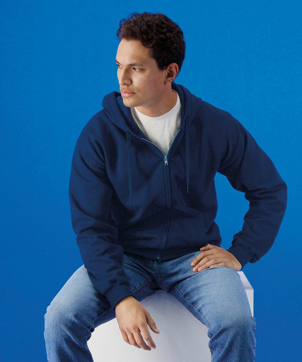 A man wearing a blue Gildan GD068 zip-up hoodie and jeans sits on a white cube against a blue background, looking slightly to the side.