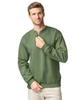 A man stands against a white background wearing the Gildan Softstyle midweight adult fleece 1/4-zip sweatshirt in green with light-coloured trousers, holding the zip with one hand and resting his other hand near his waist.