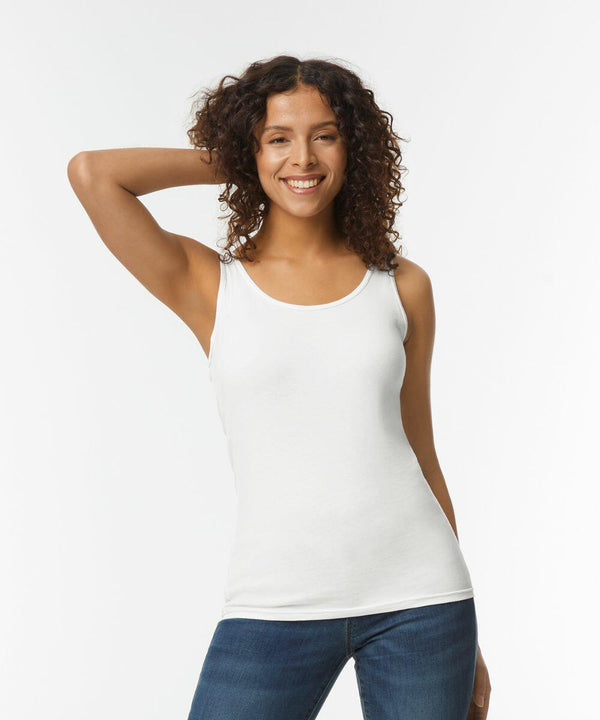 A person with curly hair smiles while posing in a Gildan GD077 white sleeveless top and blue jeans against a plain background.